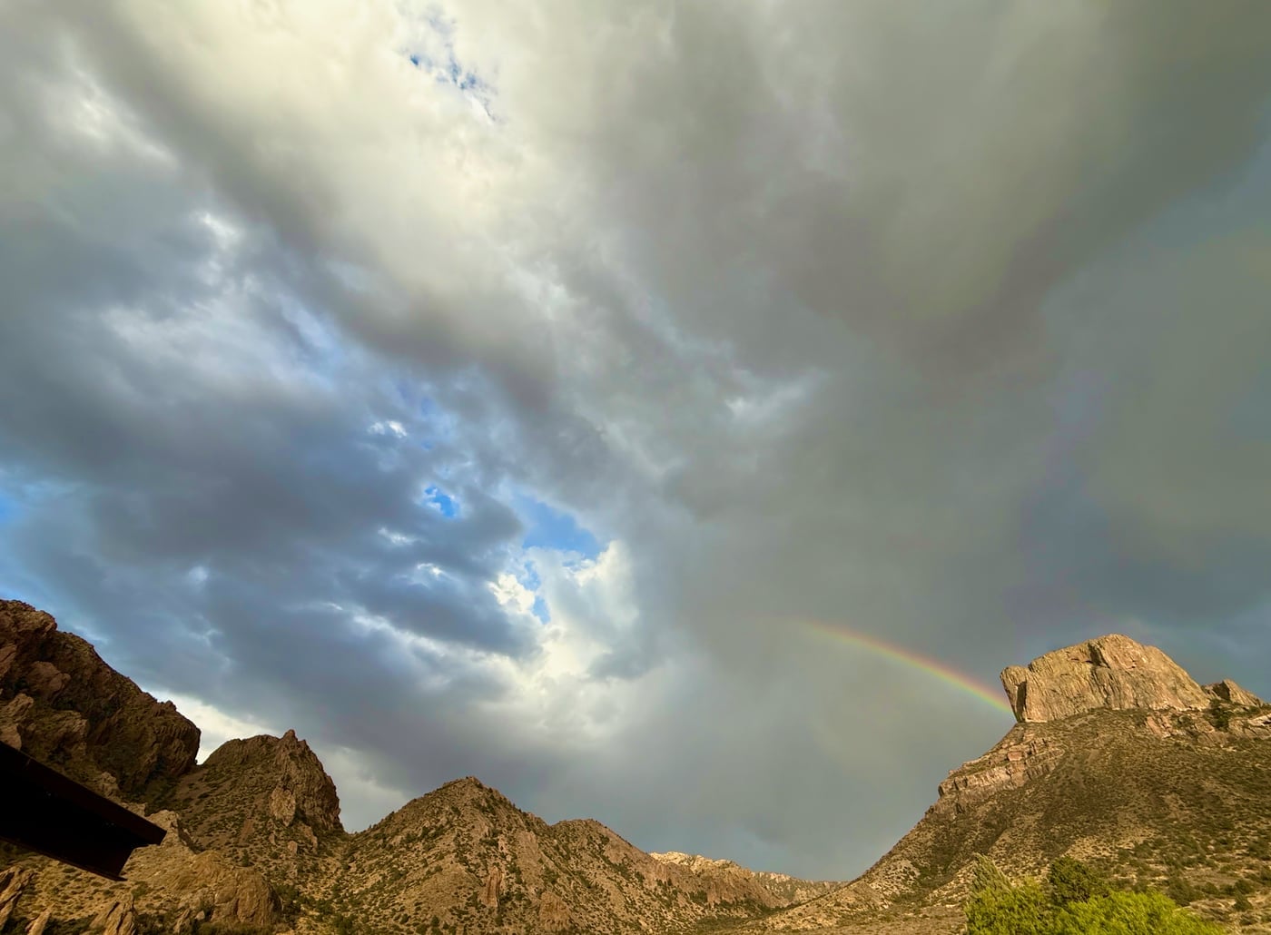 A rare desert storm passing over the Chisos Basin campground at last light, a vivid rainbow arched against the dark mountain wall, Big Bend National Park