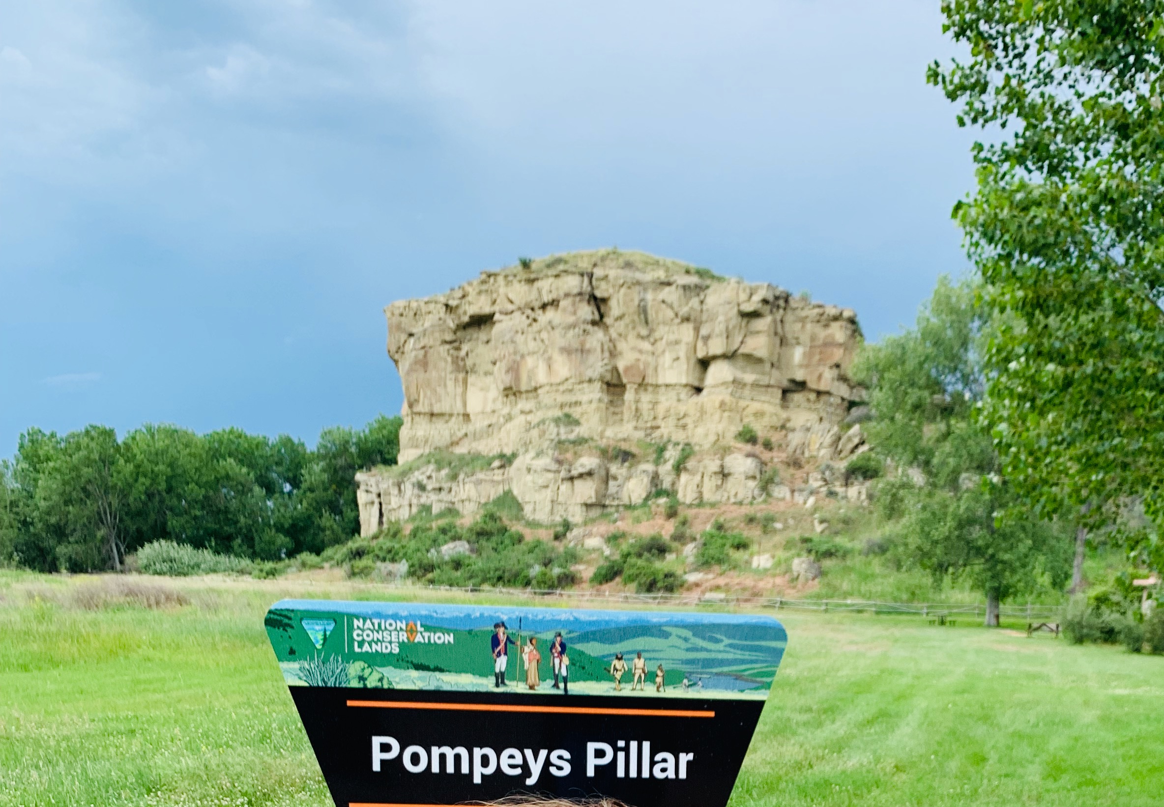 The Pompeys Pillar trailhead sign, illustrated with the Lewis and Clark expedition and Native peoples together against the landscape, with the pillar rising behind it