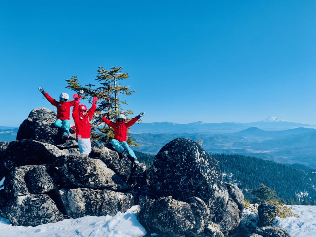 Ski kids on Mount Ashland raising their arms in the air on top of big grey boulders with a view of the Cascades and Mount Shasta in the background